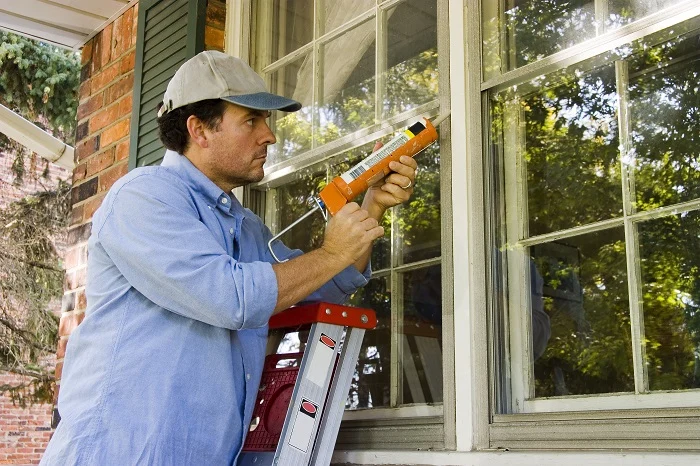 man in white hat and blue shirt caulking exterior of window while on a ladder