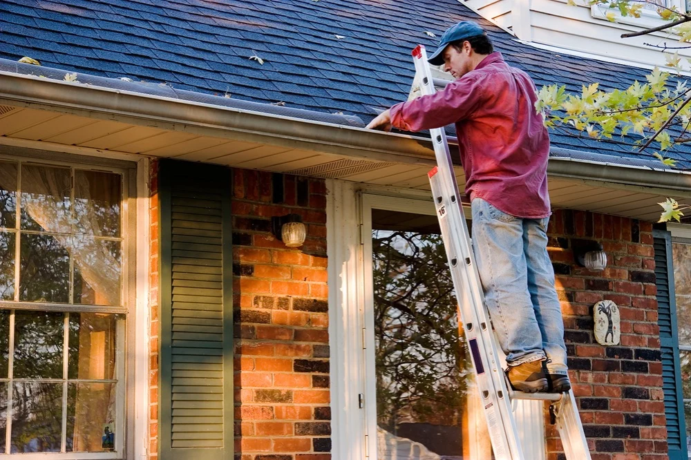 man in red shirt and blue hat on a ladder cleaning out gutters