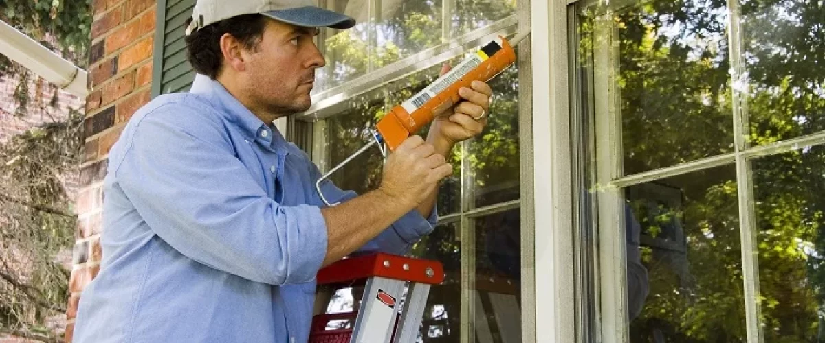 man in white hat and blue shirt caulking exterior of window while on a ladder