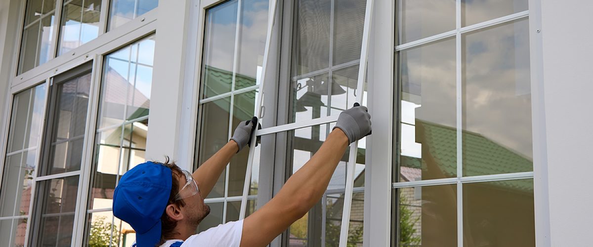 Professional window installer outside the building installs a protective insect net on large plastic window. Working man holds a mosquito net in his hands, compares the size with window