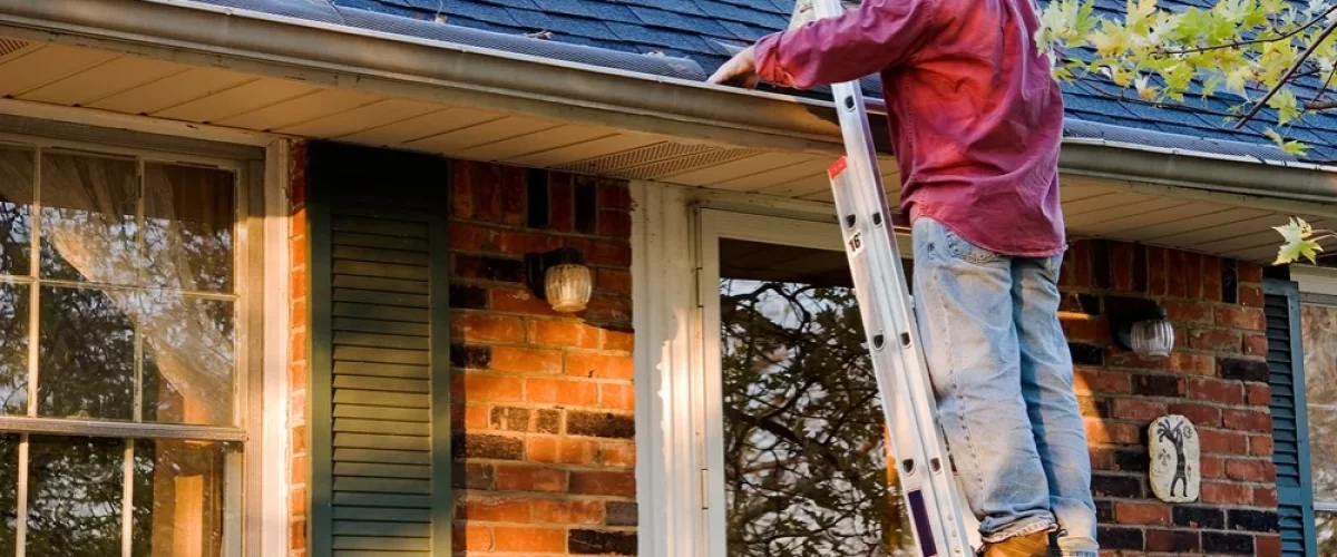 man in red shirt and blue hat on a ladder cleaning out gutters